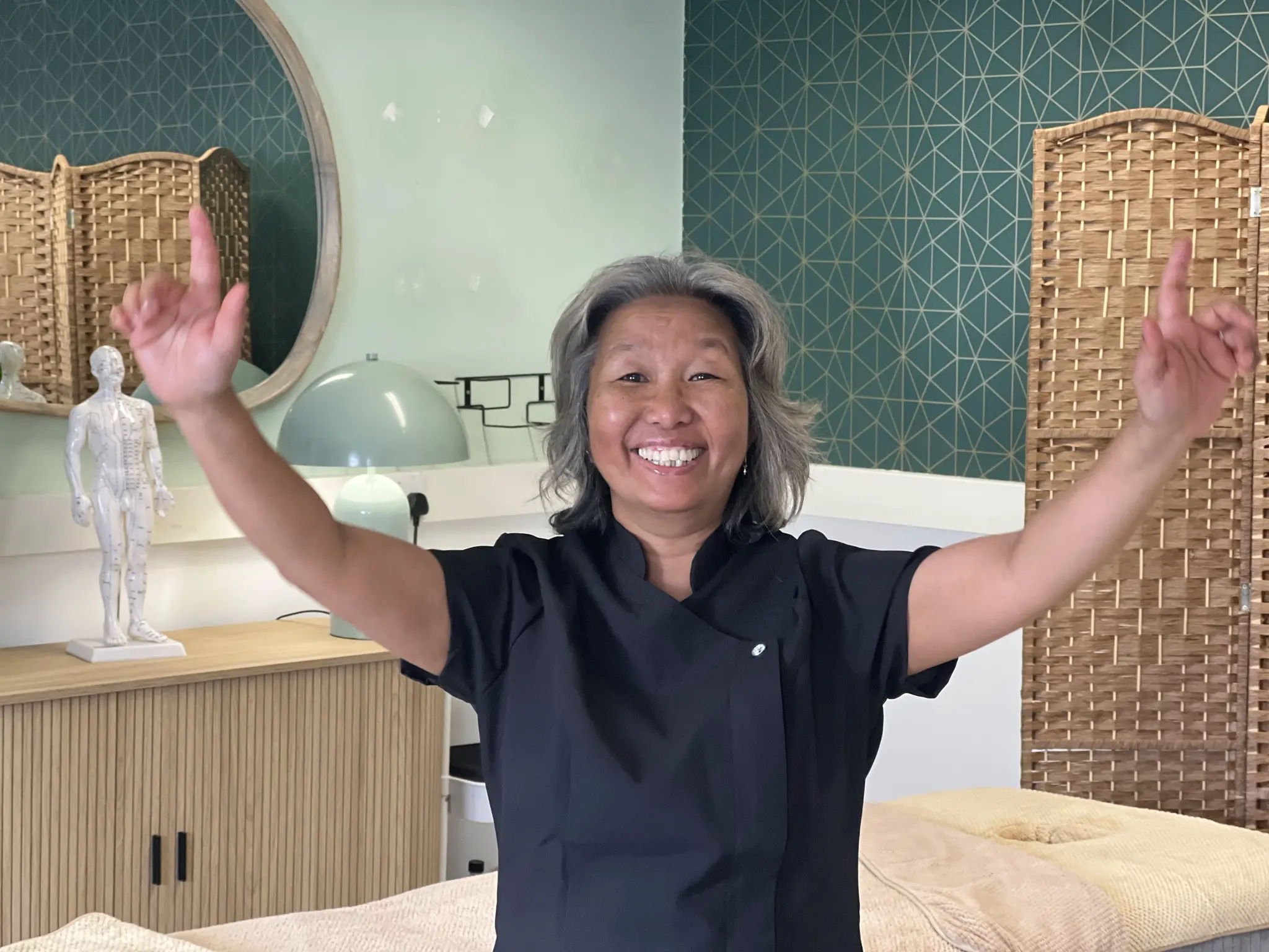 Shi Shi with both arms raised and a big smile in her treatment room, beside the anatomy figure and woven screen