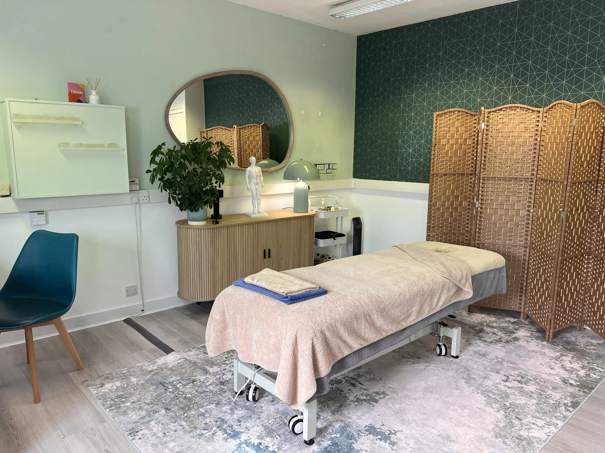 The treatment room: massage bed with a peach cover, woven screens, a round mirror and a teal chair