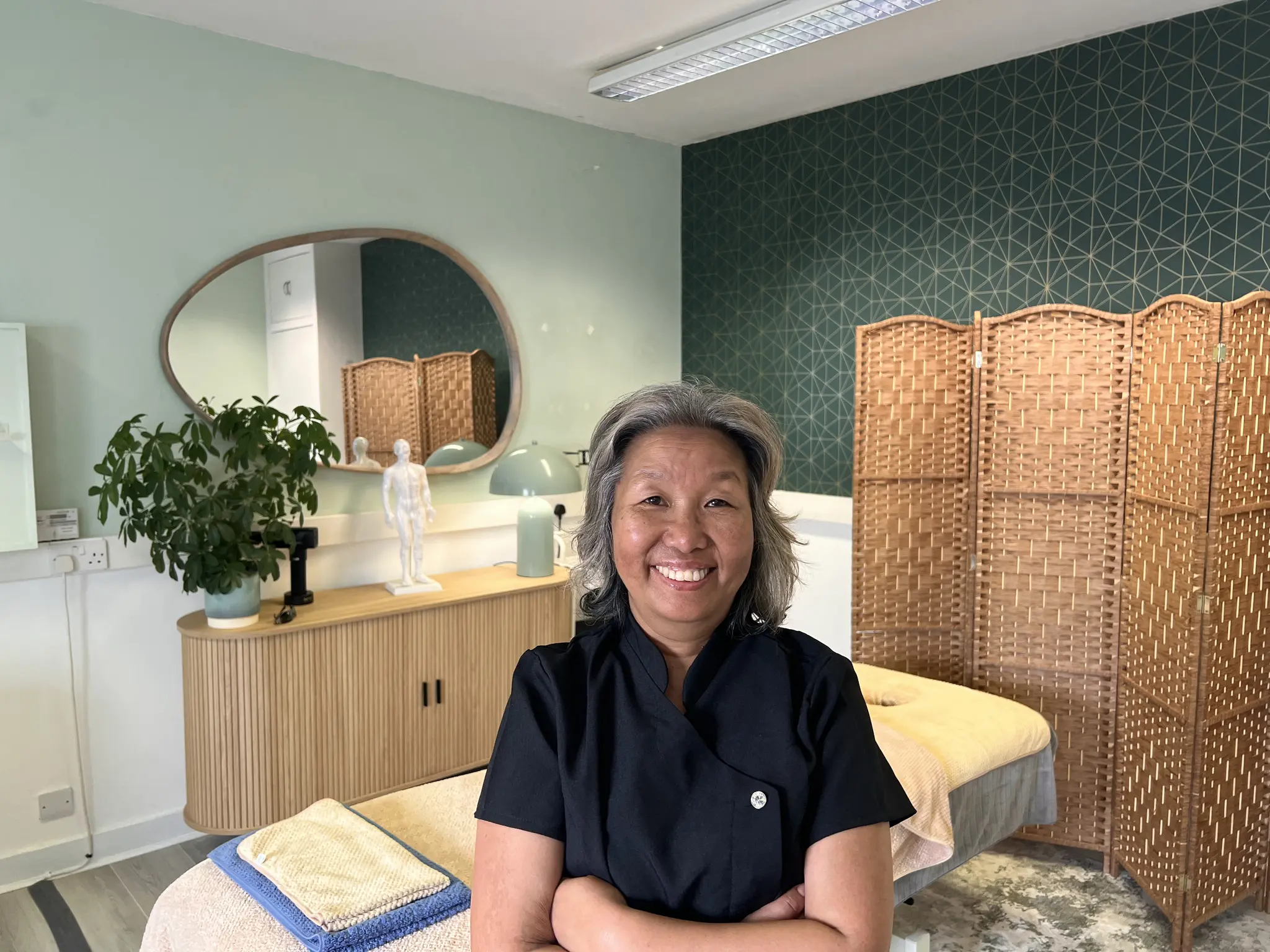 Shi Shi smiling with arms folded in her calm treatment room in Stornoway, with the massage bed and woven screens behind her