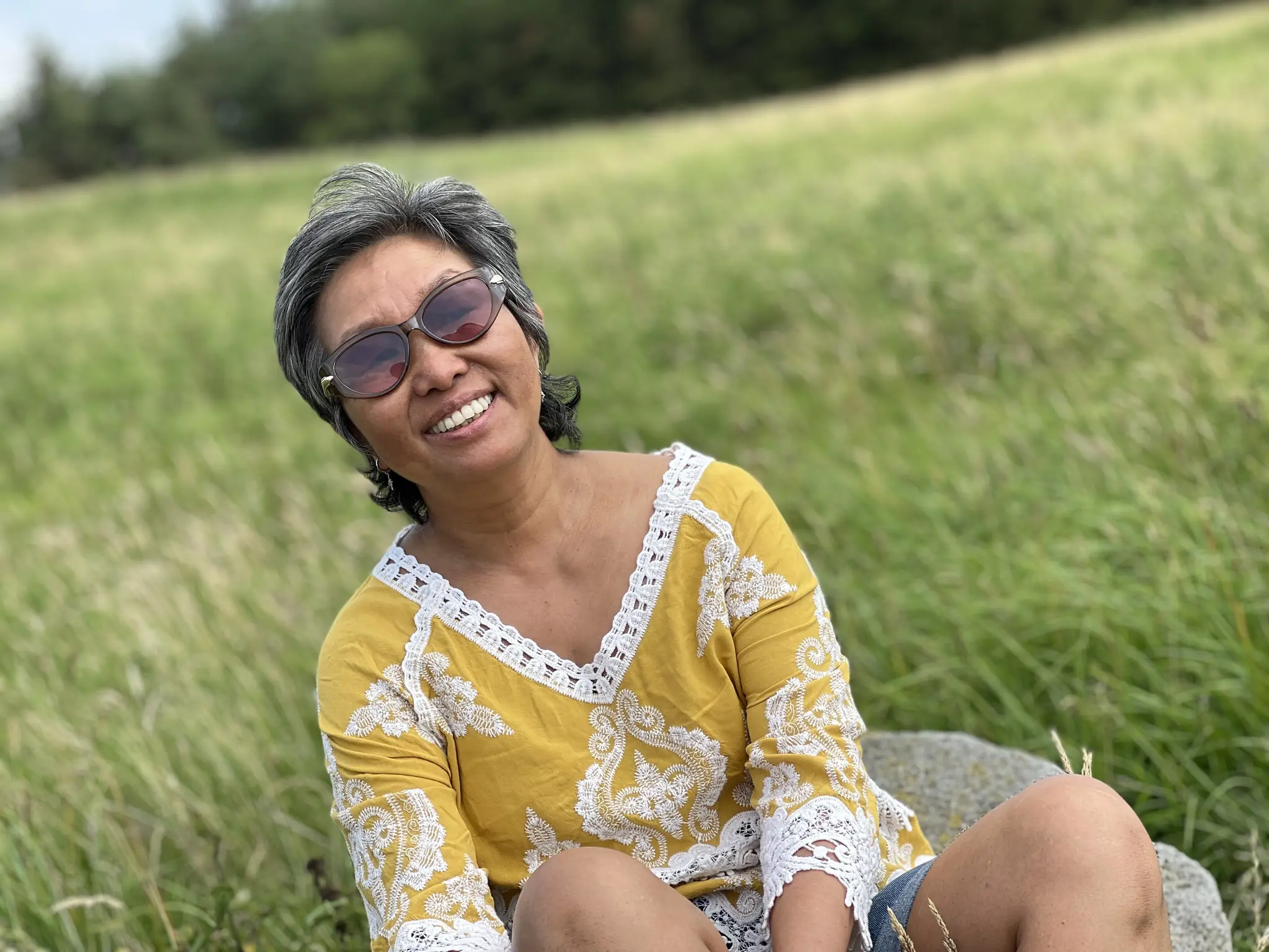 Shi Shi sitting in long grass on a sunny day, smiling, wearing sunglasses and a yellow patterned top